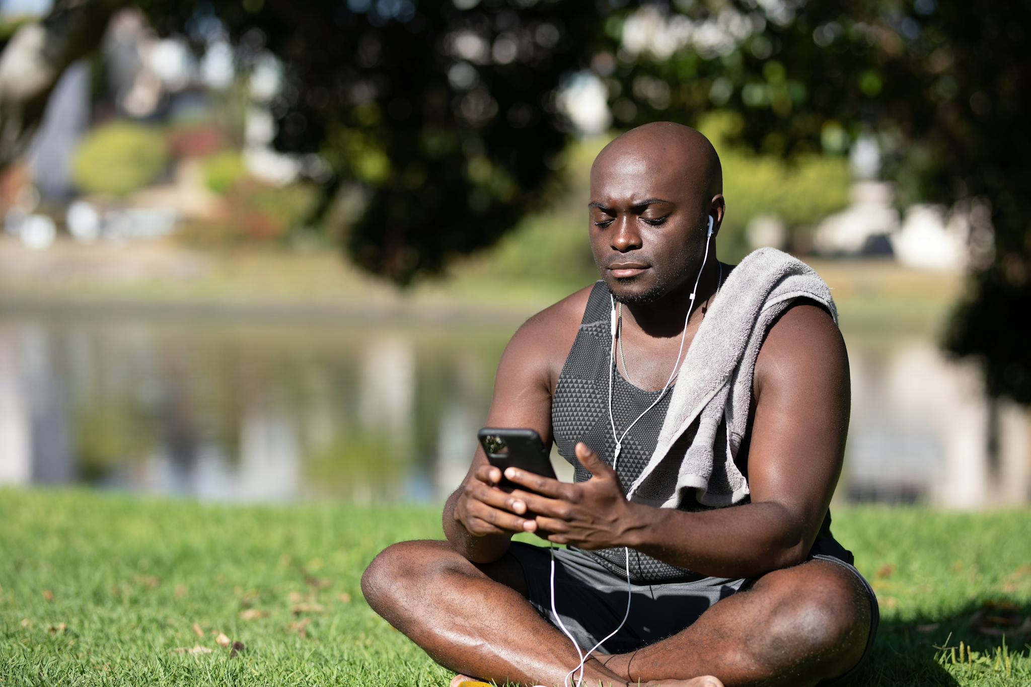Bald man sitting in park using smartphone with earphones on a sunny day, showcasing a moment of leisure.